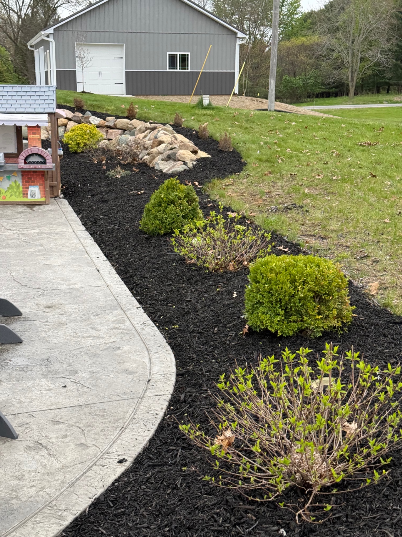 Sweeping black mulch bed with boxwoods and stacked-stone accent along stamped concrete drive